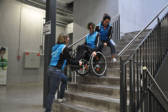 Zwei Helferinnen üben mit einer Person im Rollstuhl auf der Treppe.