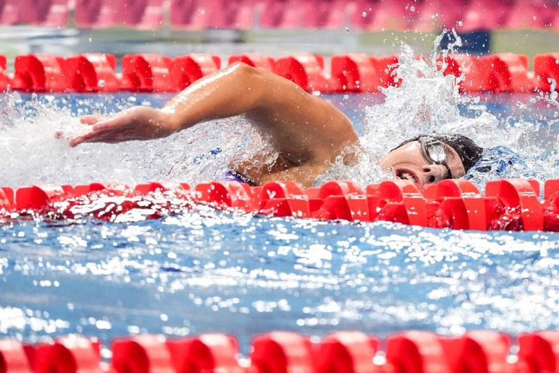 Ein Schwimmer, der eine Schwimmbrille und eine Badekappe trägt, nimmt an einem Freistilrennen teil und verursacht dabei Spritzer im Wasser, mit leuchtend roten Bahntrennern auf beiden Seiten.