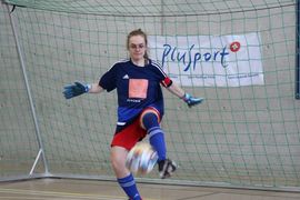 Eine Hallenfußballspielerin in einem blauen Trikot, roten Shorts und Handschuhen kickt einen Ball vor einem Netz in einer Sporthalle. Im Hintergrund ist ein Banner mit der Aufschrift PluSport zu sehen.
