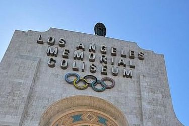Die Menschen betreten das Los Angeles Memorial Coliseum unter einem großen Torbogen, der mit den olympischen Ringen und eingravierten Schriftzügen geschmückt ist, während über ihnen ein strahlend blauer Himmel zu sehen ist.