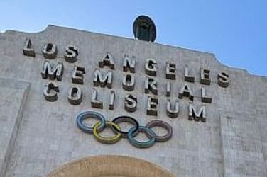 Die Menschen betreten das Los Angeles Memorial Coliseum unter einem großen Torbogen, der mit den olympischen Ringen und eingravierten Schriftzügen geschmückt ist, während über ihnen ein strahlend blauer Himmel zu sehen ist.