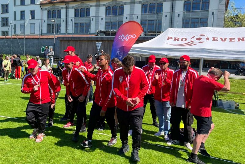 Eine Gruppe von Männern in passenden roten Trainingsanzügen und Mützen feiert mit Medaillen um den Hals auf einer Wiese, in der Nähe eines weißen Zeltes und eines Veranstaltungsbanners, mit Menschen und Gebäuden im Hintergrund.