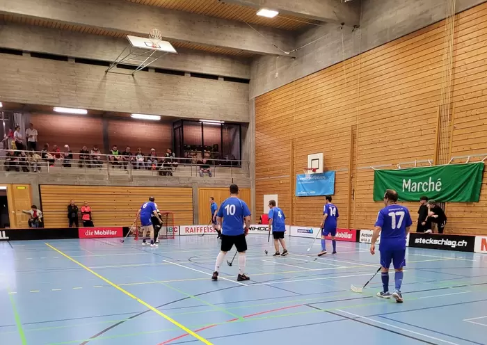 Ein Unihockeyspiel in einer Sporthalle. Spieler in blauen Trikots spielen auf einem hellblauen Spielfeld. Zuschauer beobachten das Spiel von der Tribüne im Hintergrund. Grüne und rote Banner hängen an der Holzwand.
