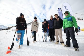 Eine Gruppe von sechs Personen, darunter auch Personen mit Beinprothesen und Krücken, steht gemeinsam auf einer verschneiten Piste. Sie tragen Winterkleidung und lächeln, im Hintergrund sind Skibanner und verschneite Berge zu sehen.