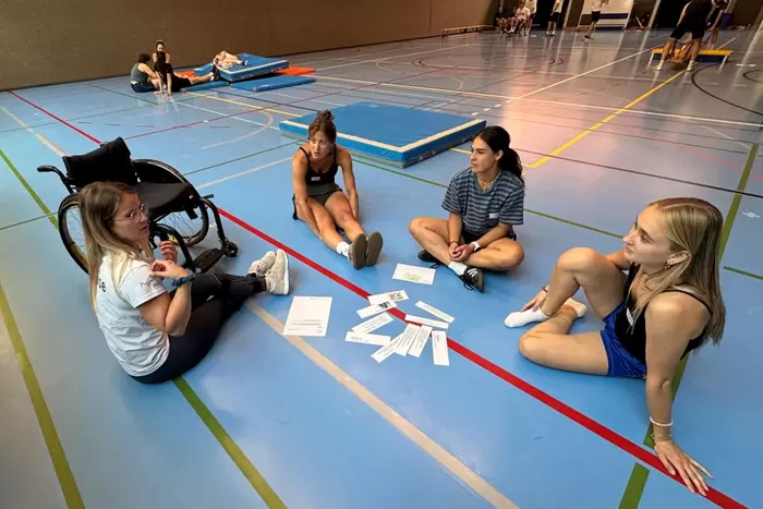 Eine Gruppe von Kursteilnehmenden sitzt auf dem Turnhallenboden und besprechen eine Übung.