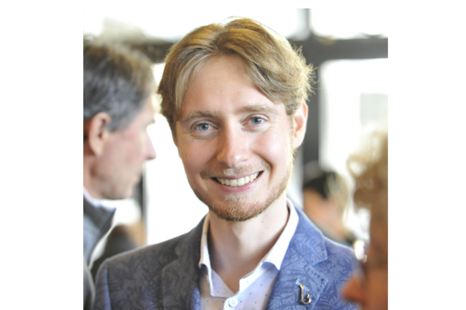 Ein lächelnder Mann mit hellbraunen Haaren und einem kurzen Bart trägt einen blau gemusterten Blazer und ein weißes Hemd und steht in einem Raum, während im Hintergrund verschwommene Menschen zu sehen sind.