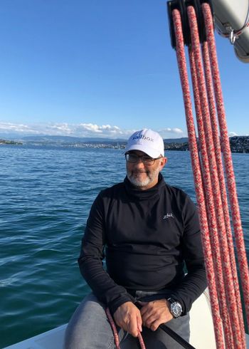 Ein Mann mit Brille, weißer Mütze und schwarzem Rollkragenpullover sitzt auf einem Segelboot und lächelt. In der Nähe hängen rote Taue, und im Hintergrund ist ruhiges blaues Wasser mit entfernten Küsten und Bergen zu sehen.