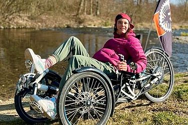 Eine Person in einer rosa Jacke und einer Mütze entspannt sich auf einem Liegerad am Flussufer. Am Liegerad ist eine schwarz-orangefarbene Fahne befestigt. Im Hintergrund sind Bäume und der blaue Himmel zu sehen.
