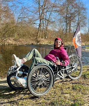 Eine Person in einer rosa Jacke und einer Mütze entspannt sich auf einem Liegerad am Flussufer. Am Liegerad ist eine schwarz-orangefarbene Fahne befestigt. Im Hintergrund sind Bäume und der blaue Himmel zu sehen.