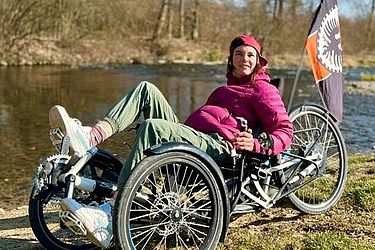 Eine Person in einer rosa Jacke und einer Mütze entspannt sich auf einem Liegerad am Flussufer. Am Liegerad ist eine schwarz-orangefarbene Fahne befestigt. Im Hintergrund sind Bäume und der blaue Himmel zu sehen.