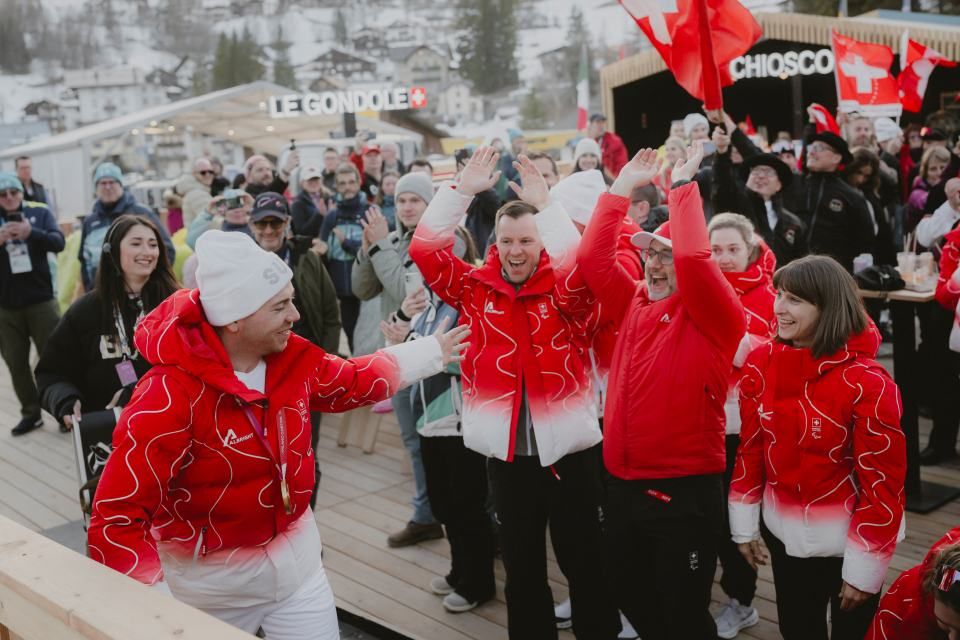Eine Gruppe von Menschen in roten und weißen Winterjacken jubelt und feiert im Freien auf einer Holzterrasse, umgeben von lächelnden Zuschauern. Im Hintergrund sind verschneite Berge und Gebäude im Chalet-Stil zu sehen.