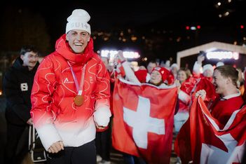 Ein lächelnder Sportler in einer rot-weißen Jacke mit einer Medaille um den Hals steht vor jubelnden Fans, die bei einer nächtlichen Freiluftveranstaltung Schweizer Fahnen halten.