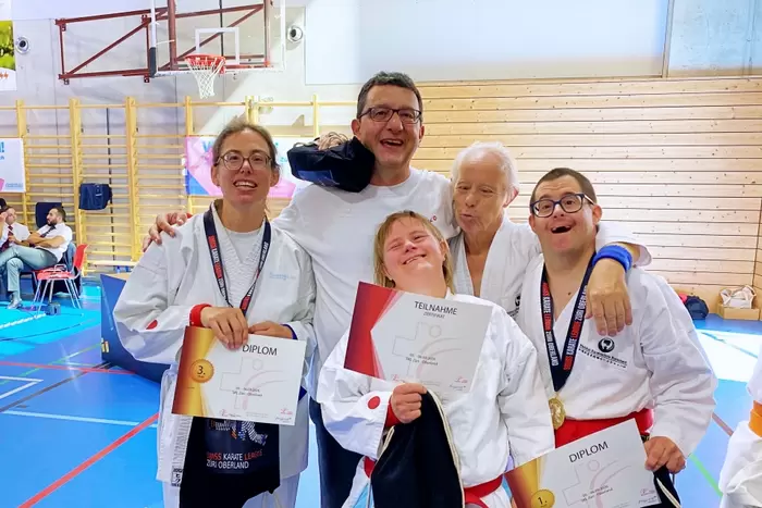 Fünf lächelnde Sportler in Kampfsportanzügen posieren gemeinsam in einer Sporthalle und halten Urkunden und Medaillen in den Händen. Sie wirken glücklich und stolz und feiern ihre Erfolge nach einem Wettkampf.