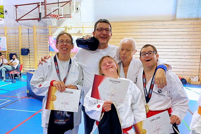 Fünf lächelnde Sportler in Kampfsportanzügen posieren gemeinsam in einer Sporthalle und halten Urkunden und Medaillen in den Händen. Sie wirken glücklich und stolz und feiern ihre Erfolge nach einem Wettkampf.