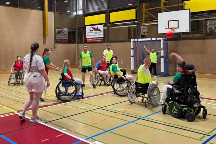 Eine Gruppe von Menschen, von denen einige im Rollstuhl sitzen, spielt in einer Sporthalle Basketball. Ein Spieler mit einer grünen Weste wirft den Ball in Richtung Korb, während andere zuschauen und am Spiel teilnehmen.