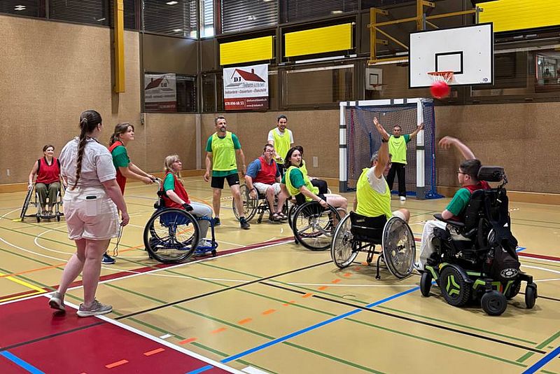 Eine Gruppe von Menschen, von denen einige im Rollstuhl sitzen, spielt in einer Sporthalle Basketball. Ein Spieler mit einer grünen Weste wirft den Ball in Richtung Korb, während andere zuschauen und am Spiel teilnehmen.