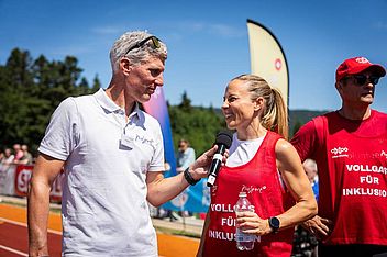 Ein Mann in einem weißen Hemd interviewt eine lächelnde Frau in einer roten Startnummer auf einer Außenbahn. Ein weiterer Mann in Rot steht daneben. Die Szene ist hell und sonnig, im Hintergrund sind Banner und Bäume zu sehen.