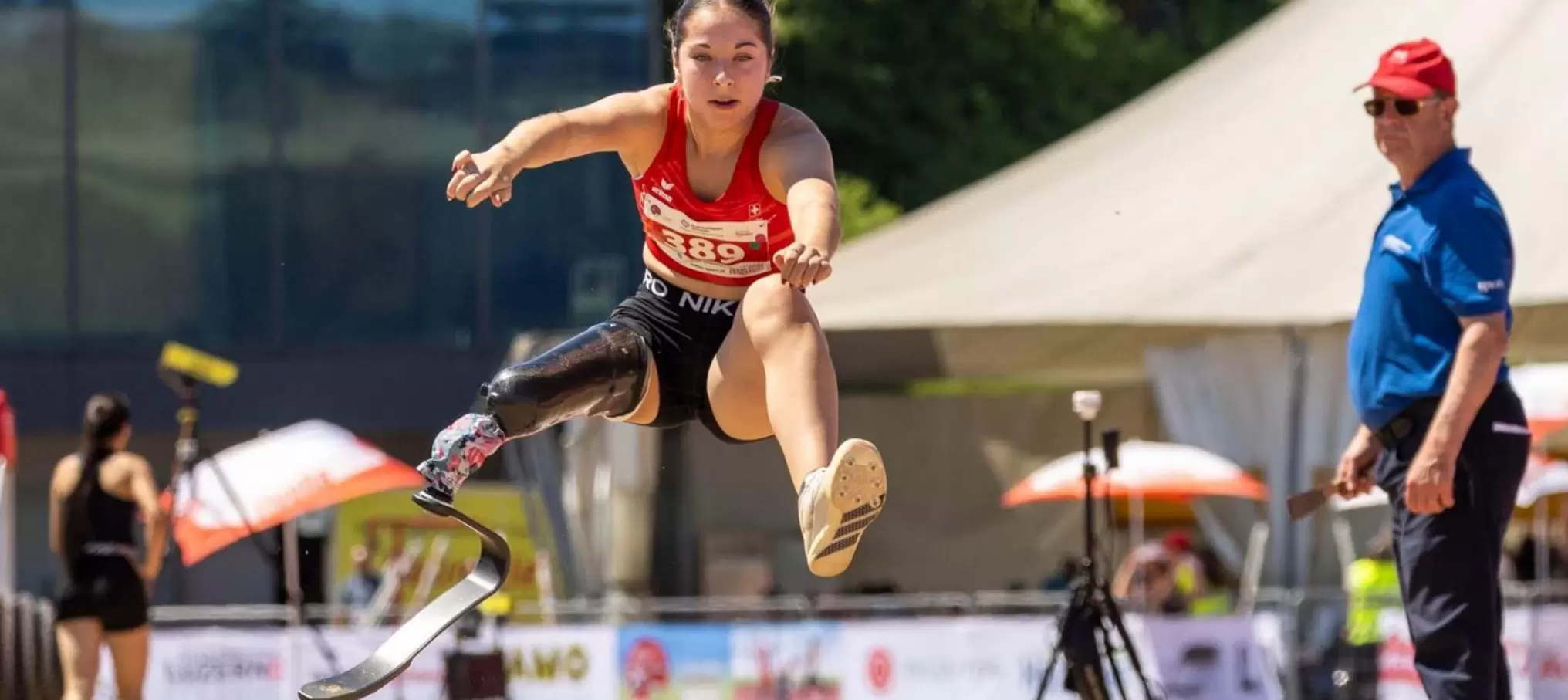 Eine Sportlerin mit einer Beinprothese befindet sich während eines Leichtathletikwettkampfs mitten im Sprung; sie trägt ein rotes Oberteil und schwarze Shorts, während im Hintergrund ein Kampfrichter zusieht.