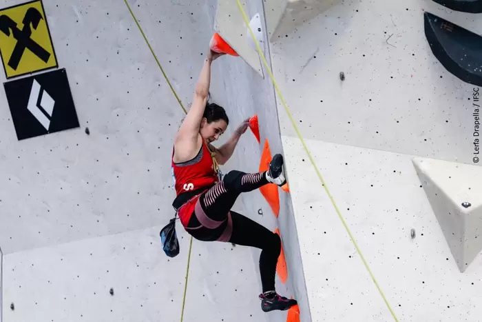 Eine Kletterin in rotem Top und schwarzen Leggings erklimmt eine Indoor-Kletterwand und klammert sich an leuchtend orangefarbenen Griffen fest. Sie wirkt konzentriert, ein Bein hochgestreckt, mit Klettergurt und Chalkbag.