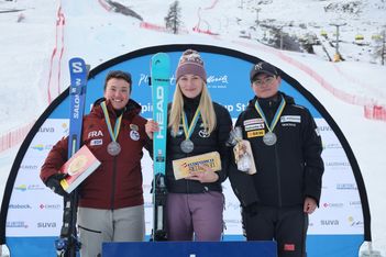Drei Skifahrerinnen stehen auf einem Podium mit Medaillen um den Hals und halten Preise in der Hand. Im Hintergrund sind verschneite Skipisten und eine Rennstrecke zu sehen. Alle drei lächeln und tragen Wintersportkleidung.