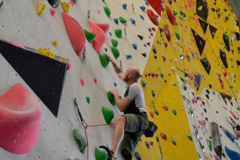 Ein Mann klettert an einer Indoor-Kletterwand und hält sich an bunten Griffen fest, während er einen Klettergurt und einen Chalkbag trägt. An der Wand gibt es verschiedene Routen, die durch Griffe in unterschiedlichen Formen und Farben gekennzeichnet sind.