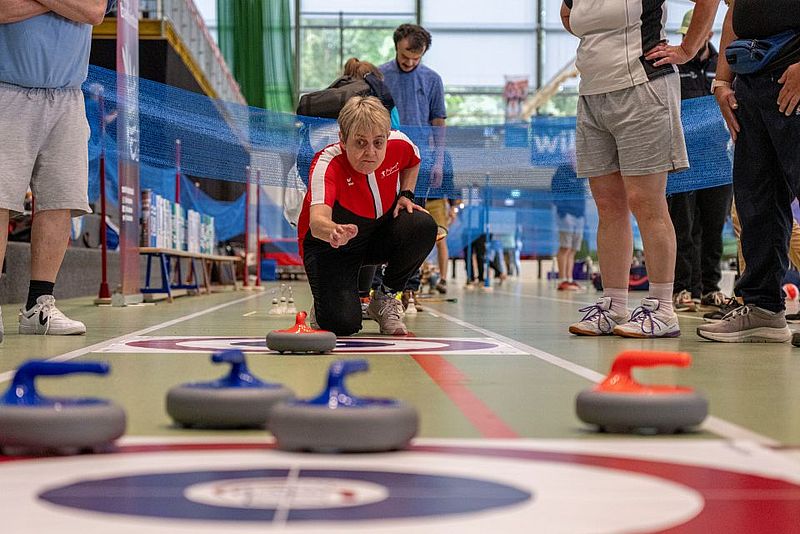 Eine Frau in einem roten Shirt hockt sich hin und macht sich bereit, einen Curlingstein auf einer Curlingbahn in der Halle zu schieben; in ihrer Nähe stehen weitere Personen, und auf dem Boden sind weitere Curlingsteine zu sehen.