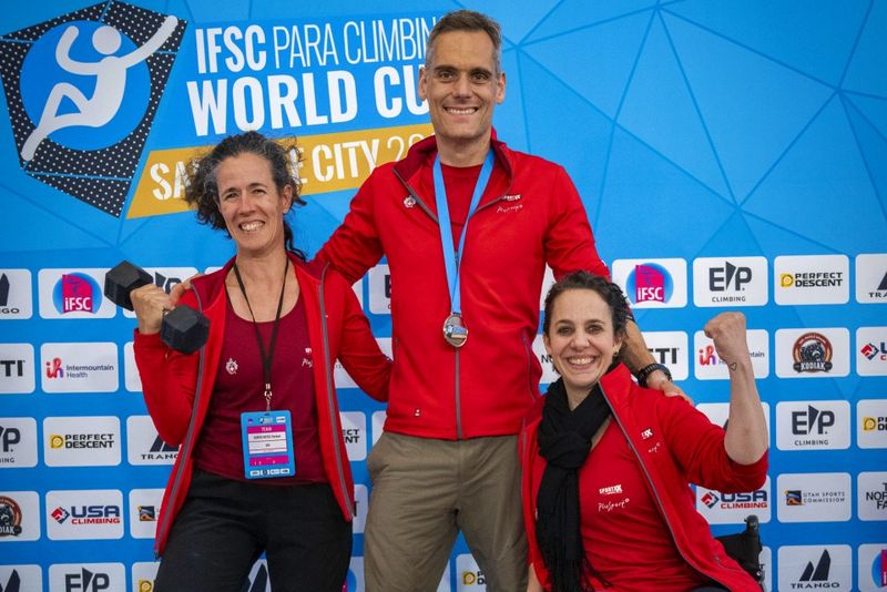 Drei lächelnde Athleten in roten Jacken posieren vor dem blauen Hintergrund des IFSC Para Climbing World Cup. Einer hält Hanteln, ein anderer trägt eine Medaille um den Hals und der dritte reckt jubelnd die Faust.