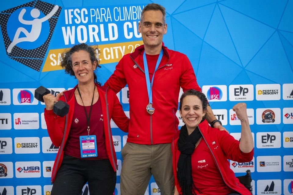 Drei lächelnde Athleten in roten Jacken posieren vor dem blauen Hintergrund des IFSC Para Climbing World Cup. Einer hält Hanteln, ein anderer trägt eine Medaille um den Hals und der dritte reckt jubelnd die Faust.