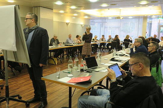 Ein Konferenzraum. Menschen sitzen an Tischen und blicken auf einen Mann, der vor einem Flipchart steht. Eine Frau steht daneben. Auf den Tischen liegen verschiedene Laptops, Papiere und Flaschen. Einige Teilnehmer konzentrieren sich auf ihre Geräte.