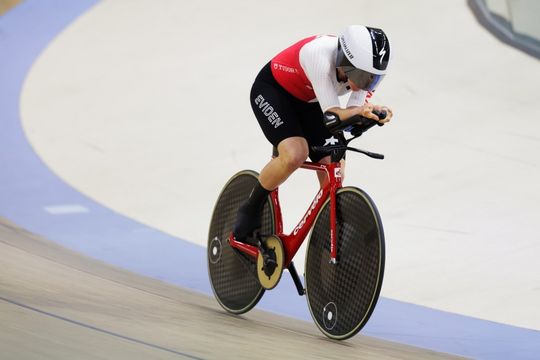 Ein Radfahrer in einem rot-weißen Trikot und schwarzen Shorts fährt auf einem Bahnrad mit Scheibenrädern in einem Velodrom, lehnt sich in einer aerodynamischen Position nach vorne und trägt einen weißen Helm.