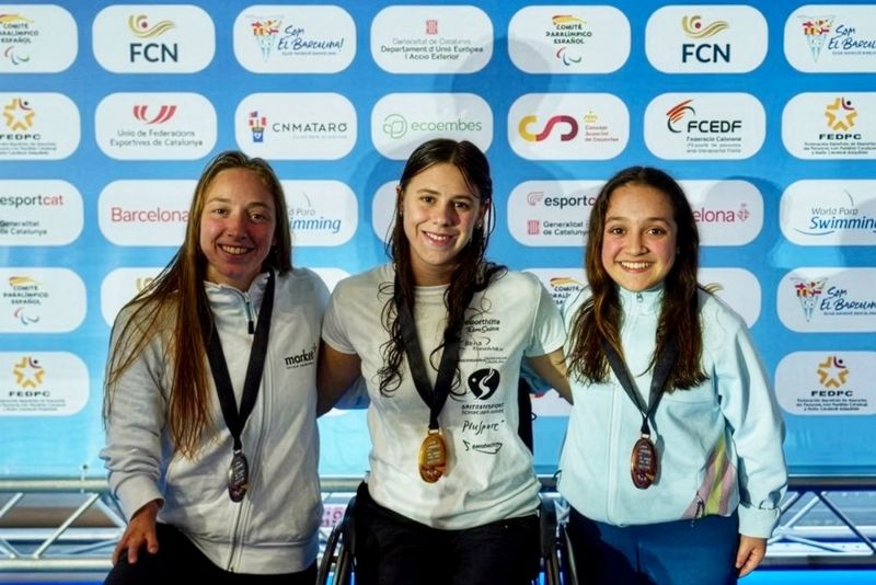 Drei Sportlerinnen mit Medaillen lächeln und posieren gemeinsam vor einem blauen, mit Logos bedeckten Hintergrund. Die mittlere Sportlerin sitzt im Rollstuhl, und alle tragen Sportjacken.