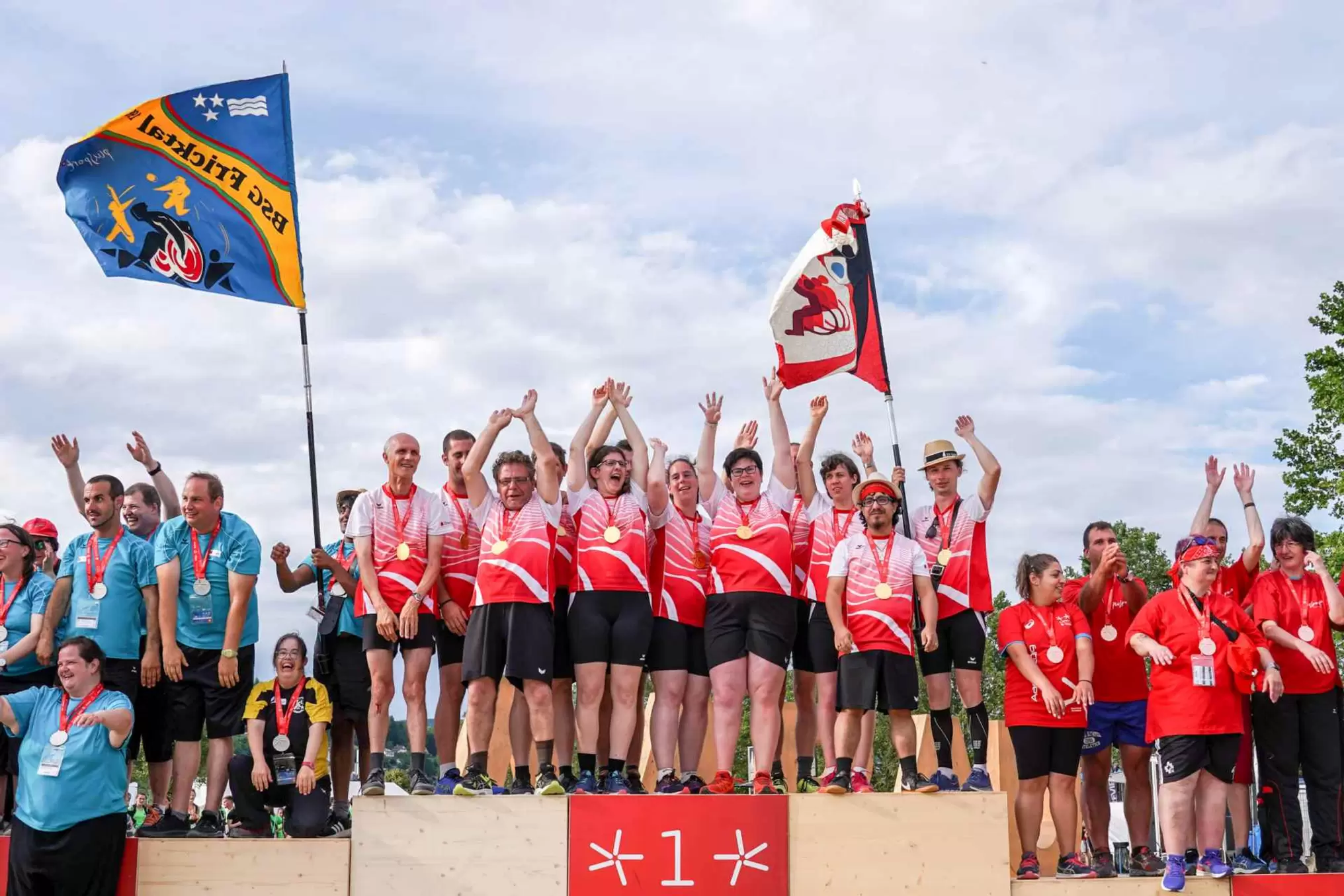 Eine große Gruppe von Menschen in passenden roten Hemden und Sportkleidung steht im Freien zusammen, viele mit erhobenen Armen und lächelnd, während andere in blauen Hemden daneben stehen und anscheinend ein Ereignis feiern oder anfeuern.