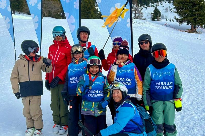 Eine Gruppe von Kindern und Erwachsenen in Skiausrüstungen und blauen "Para Ski Training"-Westen posiert gemeinsam auf einem verschneiten Berghang, im Hintergrund sind Lehrer in roten Jacken und mit Bannern zu sehen.