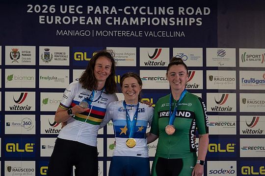 Drei Radfahrerinnen lächeln auf dem Podium, jede mit einer Medaille in der Hand, vor einer Kulisse der UEC-Para-Cycling-Straßen-Europameisterschaften 2026 in Maniago, Italien.