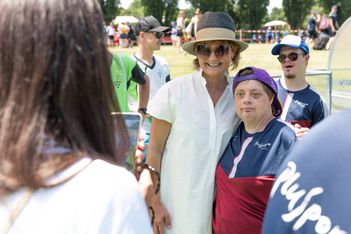 Eine Frau in einem weißen Kleid und Sonnenhut posiert mit einem lächelnden Athleten in einem roten und blauen Trikot bei einer Sportveranstaltung im Freien, während eine andere Person ihr Foto macht. Im sonnigen Hintergrund sind weitere Personen zu sehen.