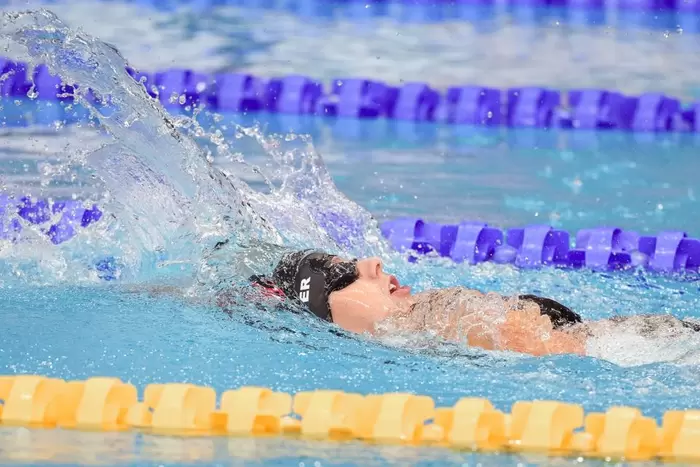 Ein Schwimmer, der eine Schwimmbrille und eine Badekappe trägt, führt in einem Schwimmbecken das Rückenschwimmen aus und erzeugt dabei einen Wasserschwall, wobei die blauen und gelben Bahnteiler sichtbar sind.