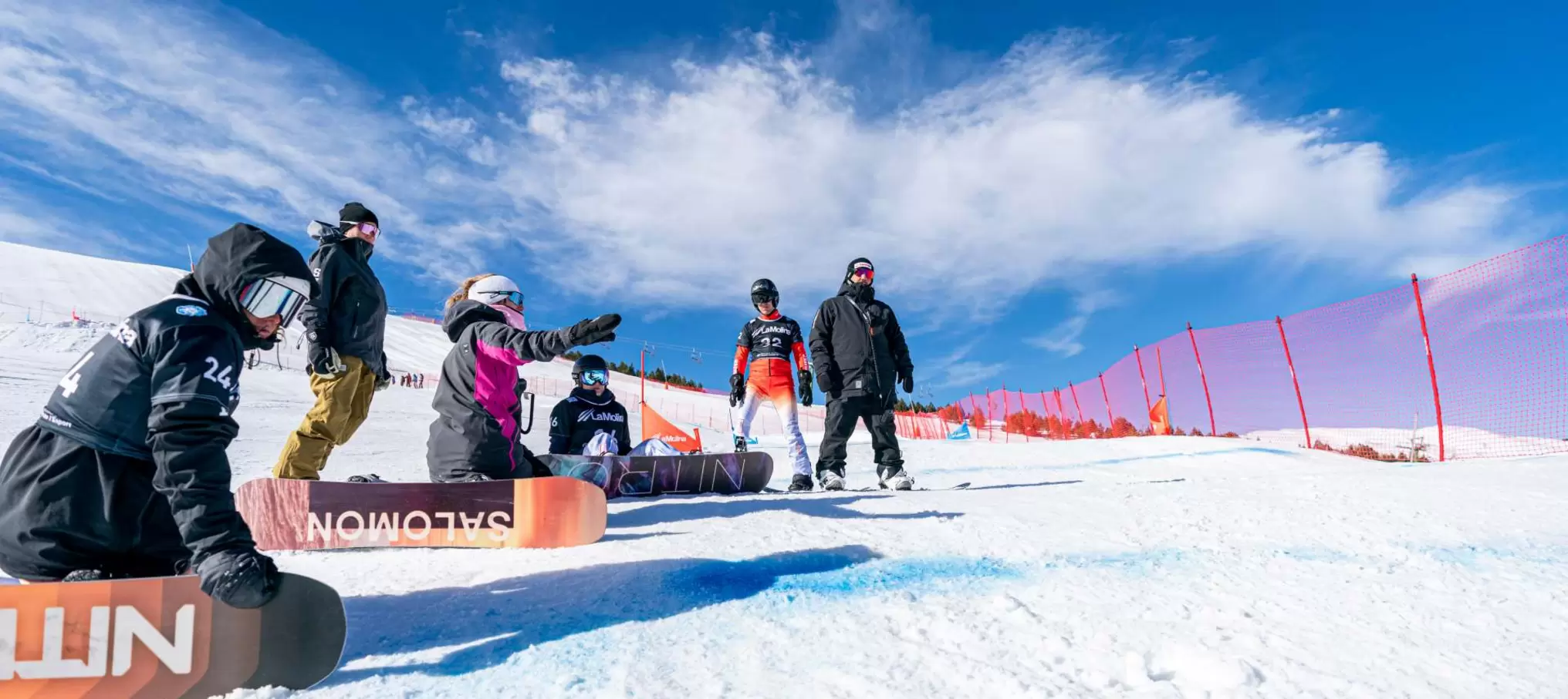 Sechs Snowboarder in voller Montur versammeln sich auf einem verschneiten Hang in der Nähe eines roten Sicherheitszauns unter blauem Himmel; eine Person kniet nieder und zeigt nach vorne, während die anderen zuschauen oder daneben stehen.