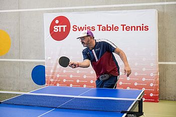 Ein Mann in Sportkleidung und mit einer Mütze spielt in einer Halle Tischtennis und konzentriert sich darauf, den Ball zu treffen. Im Hintergrund ist ein Banner des Schweizer Tischtennisverbands zu sehen.