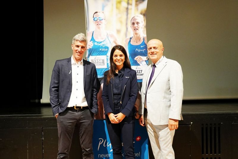 Drei Personen in Business-Kleidung stehen lächelnd vor einem Plakat, das zwei Frauen bei einem Wettlauf zeigt. Die Szene scheint bei einer Veranstaltung oder Präsentation in einem geschlossenen Raum zu spielen.