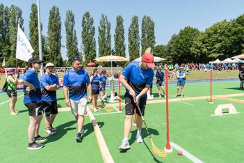 Mehrere Personen in blauen Hemden und schwarzen Shorts nehmen an einem Unihockeyturnier im Freien auf einer grünen Wiese teil, umgeben von Bäumen und Zuschauern. Eine Person mit einer roten Mütze bereitet sich darauf vor, einen Ball mit einem Schläger zu schlagen.