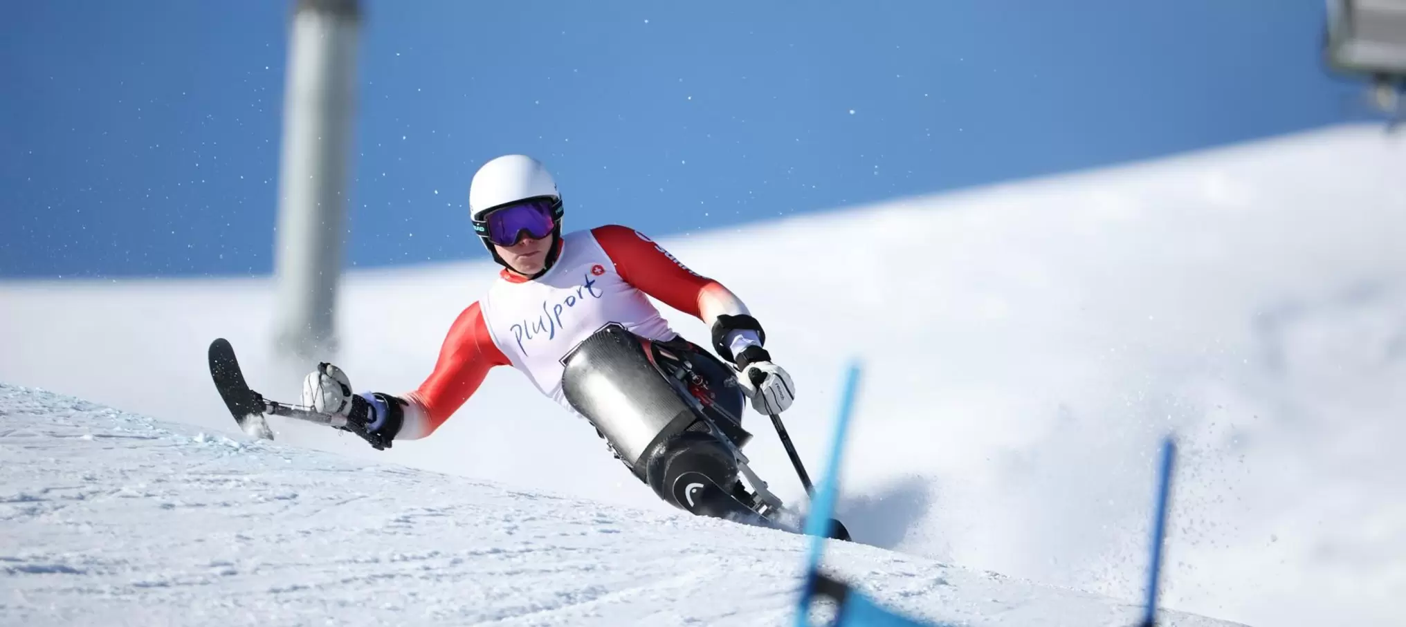 Ein Sitzskifahrer mit weißem Helm, rot-weißer Jacke und Skibrille rast bei einem Skirennen einen verschneiten Hang hinunter, wobei er unter dem strahlend blauen Himmel mit Auslegern das Gleichgewicht hält.