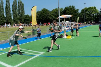 Zwei Männer in Sportkleidung tauschen bei einem Staffellauf auf einem grünen Sportplatz einen Staffelstab aus, während im Hintergrund an einem sonnigen Tag Zuschauer zusehen.