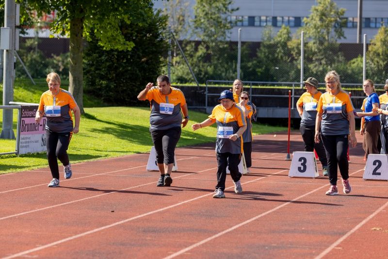 Eine Gruppe von Frauen in passenden orangefarbenen und grauen Hemden läuft auf einer Bahn und nimmt an einem Rennen teil. Sie sehen konzentriert und entschlossen aus. Im Hintergrund sind nummerierte Bahnen und grünes Gras zu sehen.