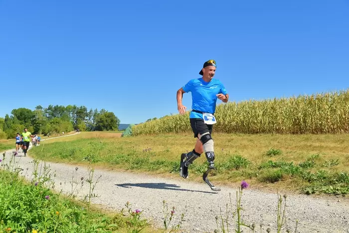 Ein Mann mit Laufschuhen läuft lächelnd auf einem ländlichen Schotterweg, umgeben von Gras und Maisfeldern, unter einem klaren blauen Himmel. Andere Läufer sind im Hintergrund zu sehen.