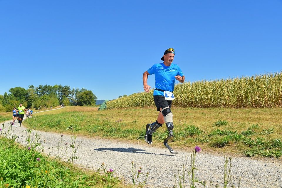Ein Mann mit Laufschuhen läuft lächelnd auf einem ländlichen Schotterweg, umgeben von Gras und Maisfeldern, unter einem klaren blauen Himmel. Andere Läufer sind im Hintergrund zu sehen.