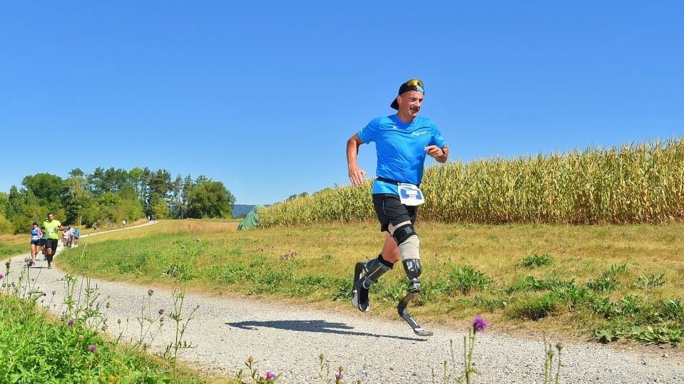 Ein Mann mit Laufschuhen läuft lächelnd auf einem ländlichen Schotterweg, umgeben von Gras und Maisfeldern, unter einem klaren blauen Himmel. Andere Läufer sind im Hintergrund zu sehen.