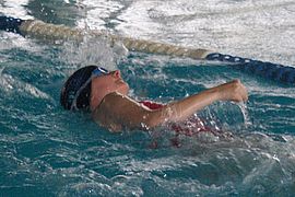 Ein Schwimmer mit Schwimmbrille und Badekappe schwimmt im Rückenschwimmstil in einem Schwimmbecken, wobei ein Arm aus dem Wasser ragt und im Hintergrund die Bahnbegrenzungen zu sehen sind.
