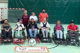 Eine Gruppe von acht Personen, einige in Elektrorollstühlen, andere stehend, posiert in einer Turnhalle neben einem großen Fußball und einem Banner mit der Aufschrift GENÈVE und dem Logo der Special Olympics.