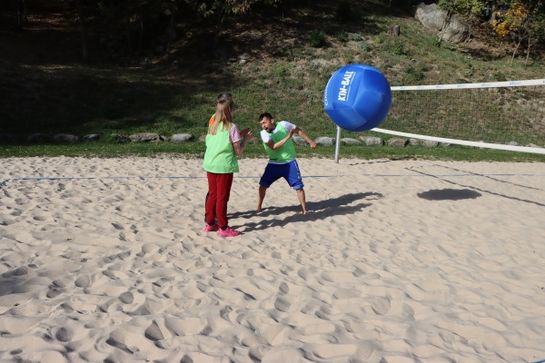 Zwei Personen, die grüne Westen tragen, spielen mit einem großen blauen Ball auf einem sandigen Platz mit einem Volleyballnetz. Eine Person ist dabei, den Ball zu schlagen, während die andere zuschaut.