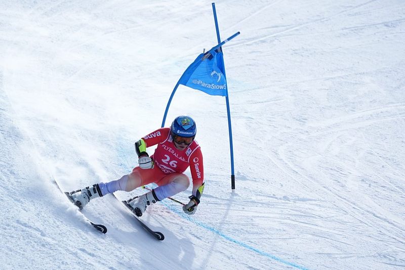 Ein Skifahrer in rotem Anzug und mit Helm rast auf einer verschneiten Piste um eine blaue Fahne herum und neigt sich bei der Kurvenfahrt stark.